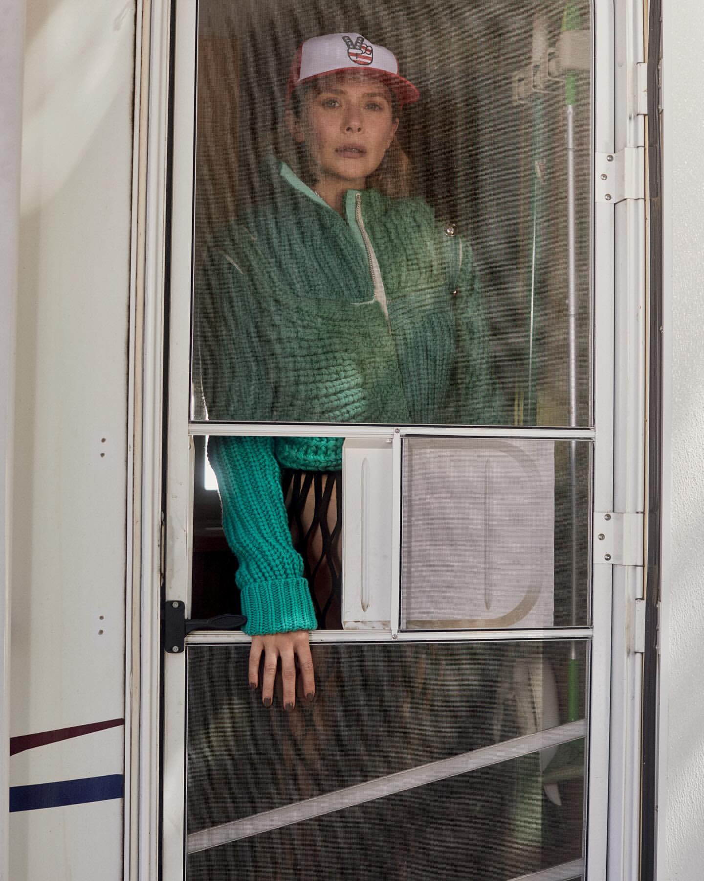 Woman in a green knit jacket and baseball cap stands in a doorway with a screen door, looking out with a neutral expression.