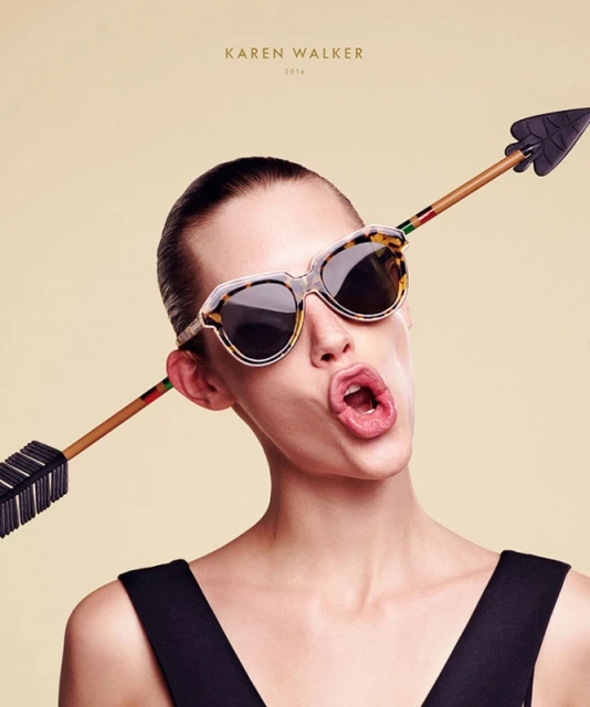 Woman poses with sunglasses pierced by arrow in fashion shot, against cream background, showcasing unusual eyewear design.