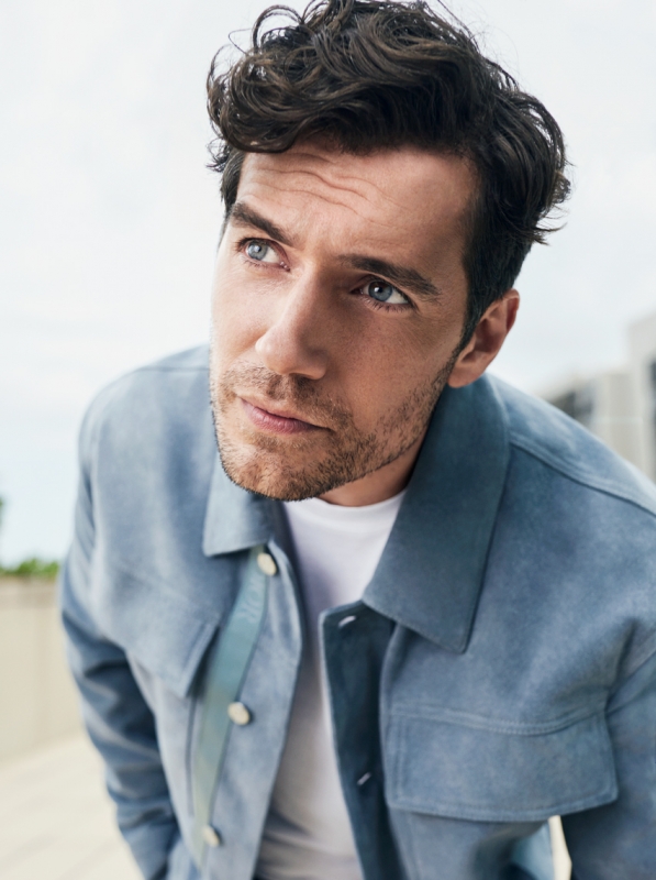 Portrait of a handsome adult male with dark hair, wearing a light blue suede jacket and white t-shirt, looking up against a bright sky.