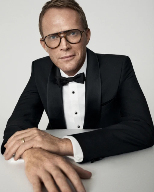 Close-up of Paul Bettany wearing glasses and a tuxedo with a bow tie, sitting at a white table against a neutral background.