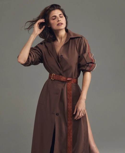 A woman in a brown trench coat with a matching belt poses indoors, her hand near her head, showcasing a blend of fashion and elegance.