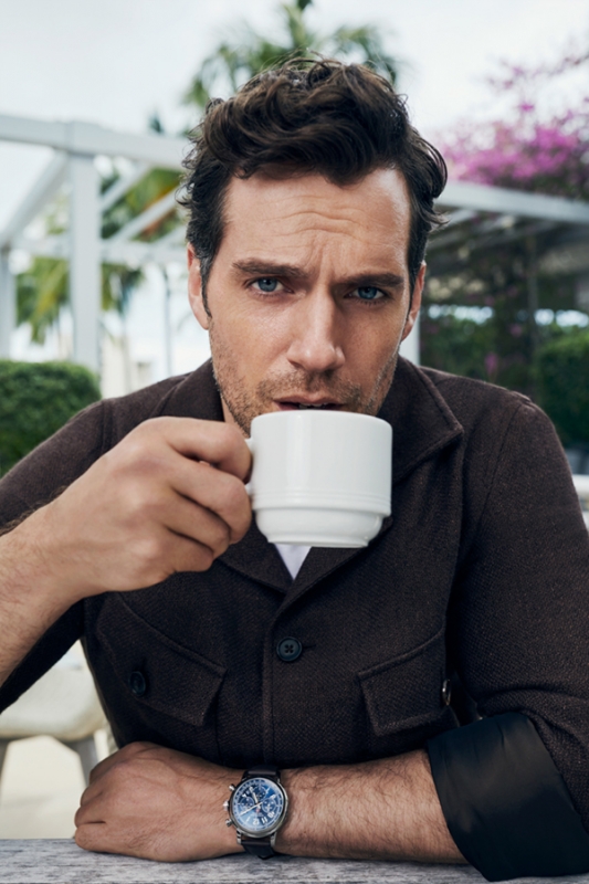 Close-up of a fair-skinned adult male drinking from a white mug outdoors, wearing a brown jacket, watch, against a floral background.