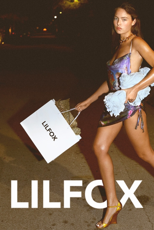 Woman walking at night holding a shopping bag and towel, wearing a patterned slip dress and wedge heels.
