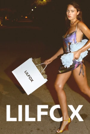 Woman walking at night holding a shopping bag and towel, wearing a patterned slip dress and wedge heels.