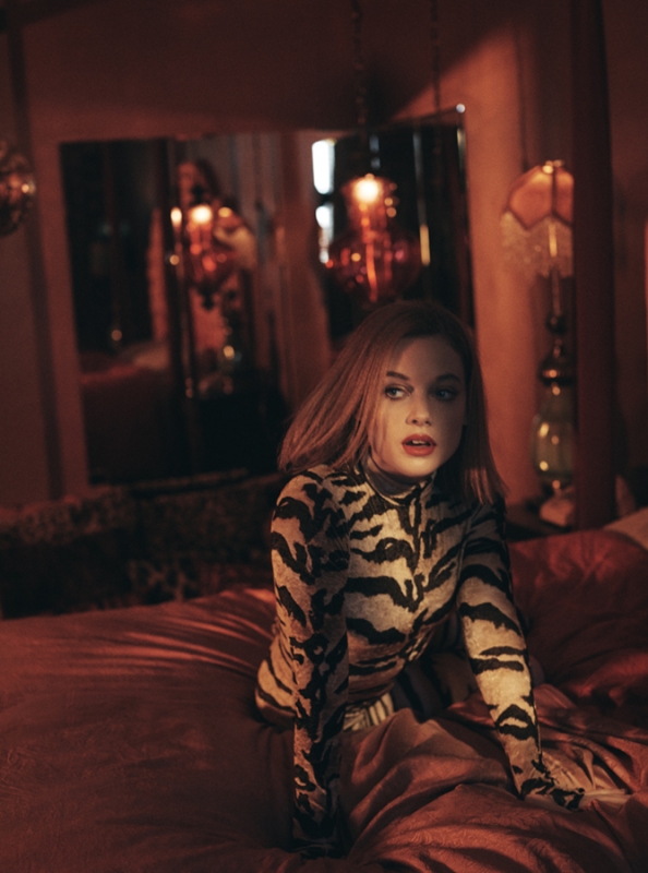 Young woman in animal print top posing on bed in dimly lit bedroom with red tones and vintage decor, looking off to the side.