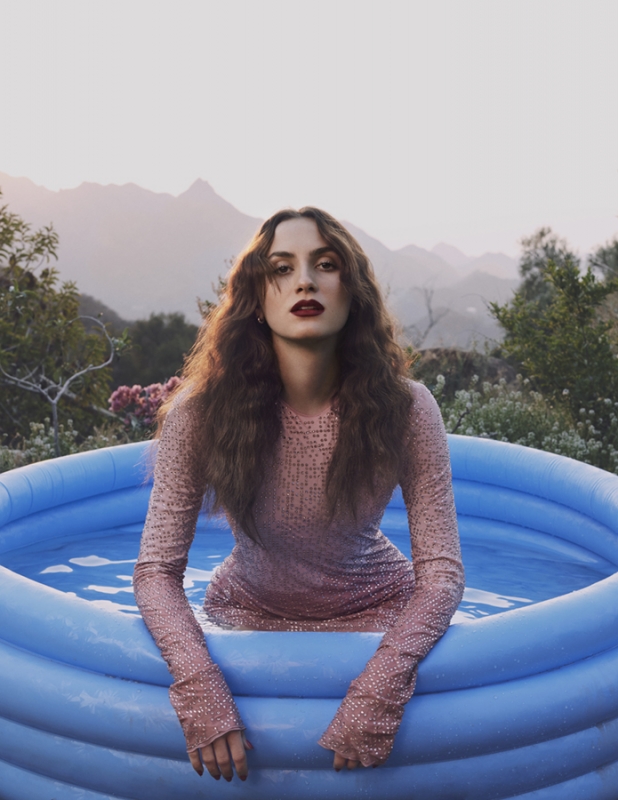 Woman in a sparkly dress sits in a blue inflatable pool with mountains and sky visible in the background in an outdoor setting.