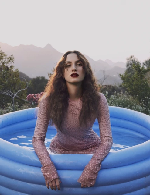 Woman in a sparkly dress sits in a blue inflatable pool with mountains and sky visible in the background in an outdoor setting.