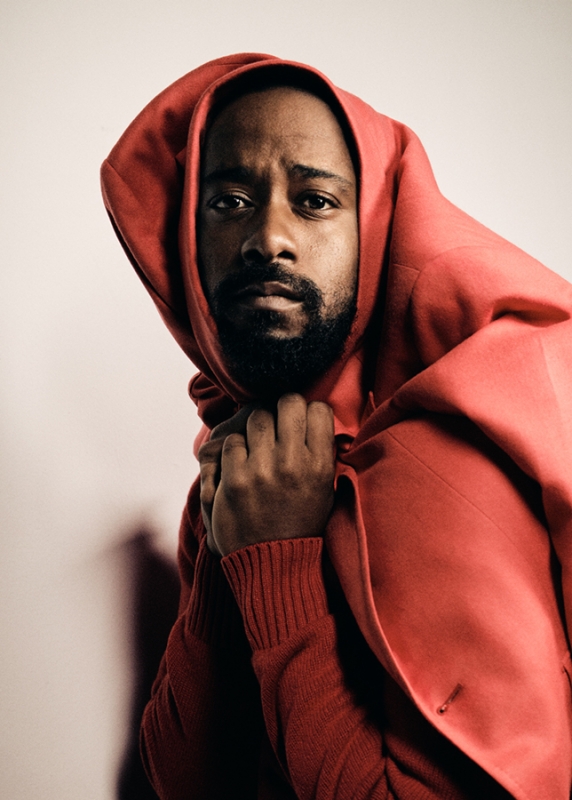 Headshot of an adult male with a beard, wearing a red hooded jacket and sweater, posing indoors against a neutral-toned background.