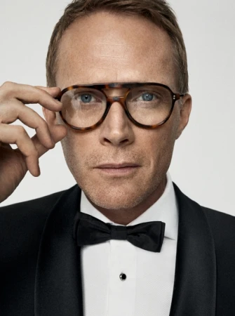 Close-up portrait of a fair-skinned adult male with short hair and glasses, wearing a black tuxedo and bow tie against a neutral background.