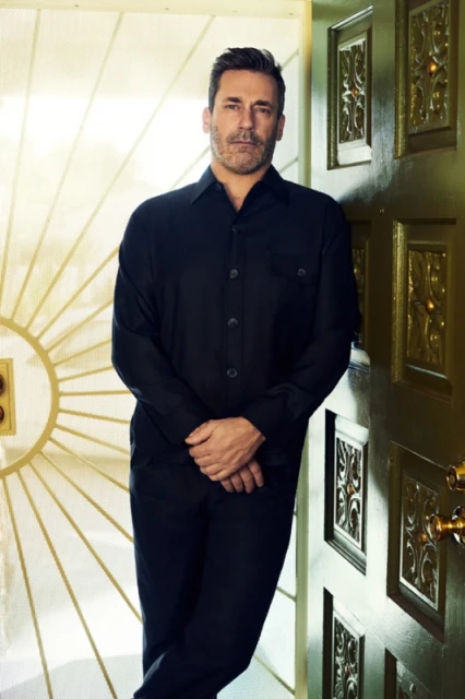 Adult male in dark button-down shirt leans against decorative green and gold wall for a portrait in an indoor setting.