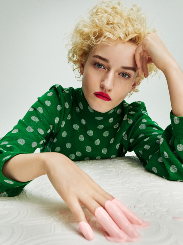 Woman with short blonde curly hair wearing a green polka dot top with her hand resting on a white patterned surface.