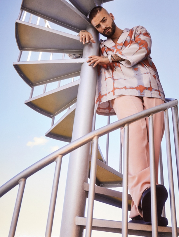 Adult male in a patterned shirt and pink pants poses on a silver spiral staircase against a bright blue sky, looking at the camera.