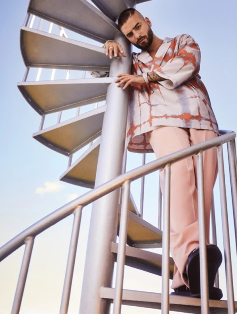 Adult male in a patterned shirt and pink pants poses on a silver spiral staircase against a bright blue sky, looking at the camera.