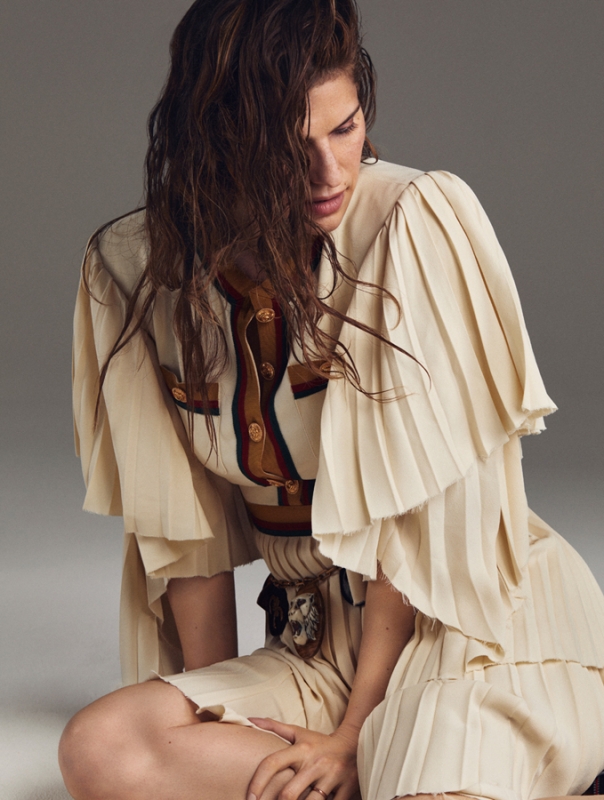 Portrait of a woman with long brown hair wearing a pleated, layered tan dress in a studio setting against a light gray backdrop.