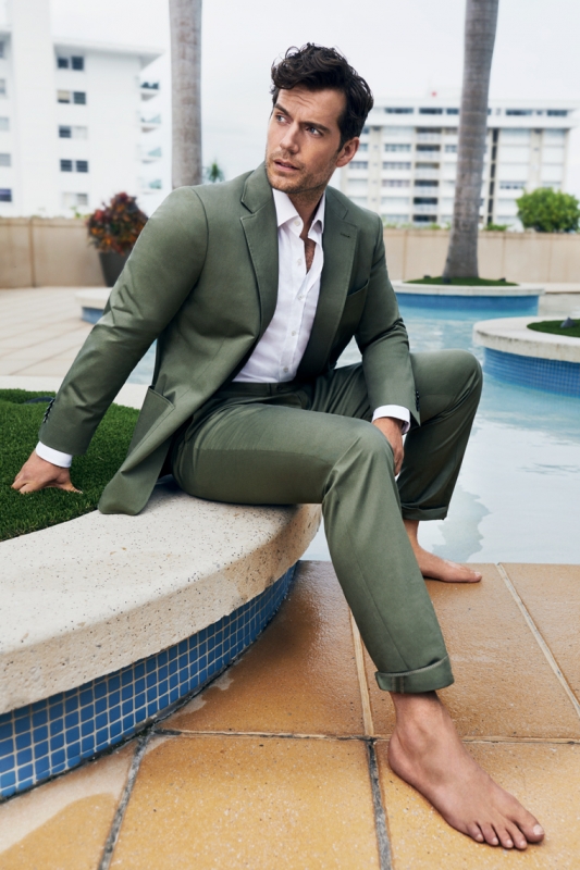 Henry Cavill poses barefoot beside a pool in an olive-green suit and open white shirt with palm trees and a building in the background.