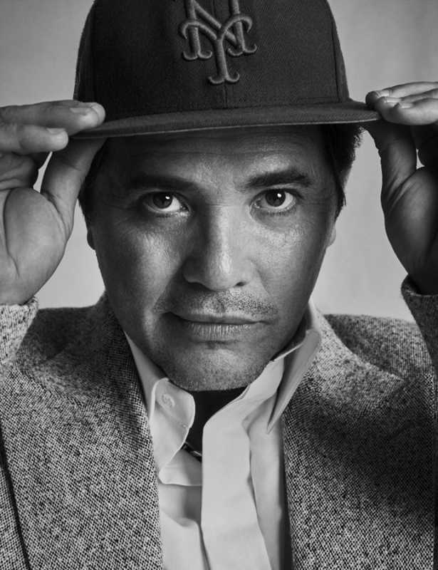 Monochrome portrait of an adult male wearing a baseball cap and grey tweed blazer looking directly at the camera with a neutral expression.