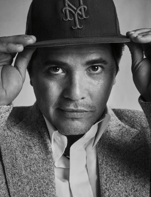 Monochrome portrait of an adult male wearing a baseball cap and grey tweed blazer looking directly at the camera with a neutral expression.