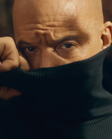 Close-up portrait of Vin Diesel pulling a dark turtleneck sweater up over his lower face with intense expression