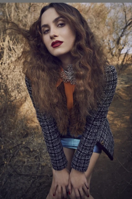 Young woman with long curly hair posing in a tweed blazer and blue shorts in a dry, natural outdoor environment with a neutral expression.