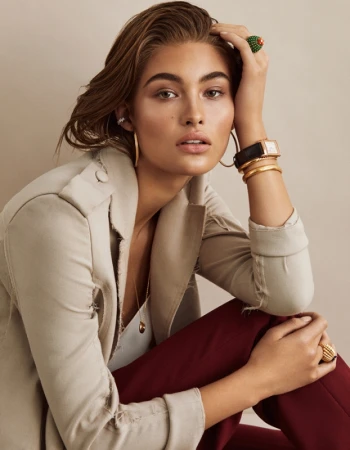 Close-up portrait of a woman in a beige jacket and burgundy trousers, wearing statement rings, earrings, and a watch.