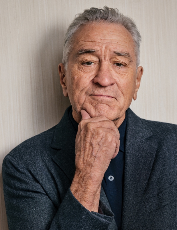 Close-up portrait of Robert De Niro in a thoughtful pose. He is wearing a dark blazer over a navy shirt with a neutral wall backdrop.