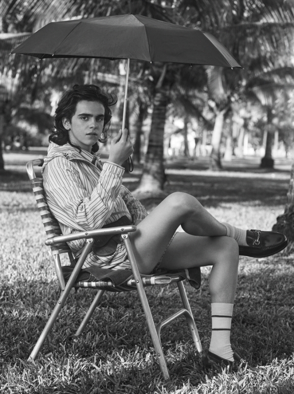 Black and white portrait of a slender adult male model relaxing on an aluminum chair outdoors with an umbrella, in a grassy landscape.