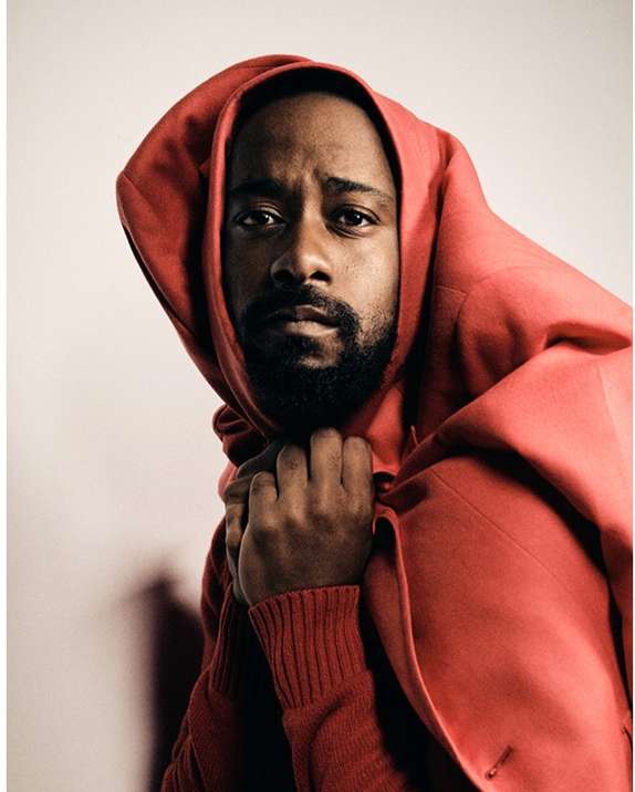 Portrait of African-American man in bright red hooded coat against neutral backdrop, captured in a medium shot with soft lighting.