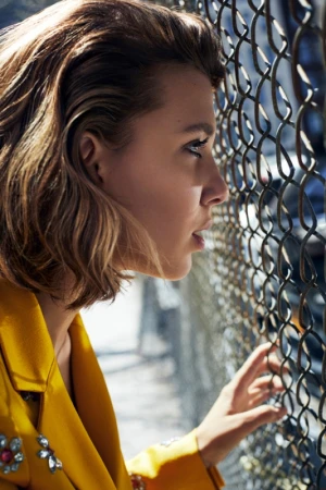Profile portrait of a woman in a yellow jacket looking through a chain-link fence outdoors.