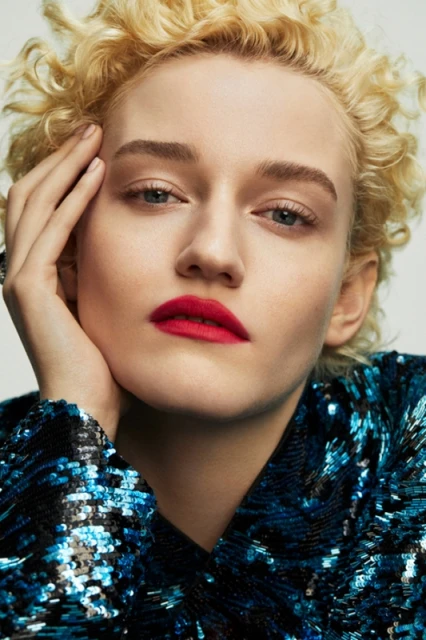 Close-up studio portrait of a young adult female with curly blonde hair, red lipstick, and a sequined jacket on a white background.
