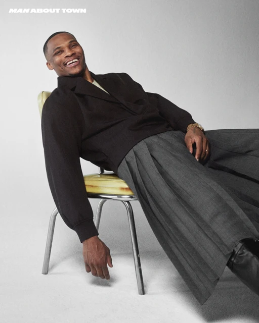Man in black jacket and grey pants smiles while seated on a vintage yellow chair in a light studio setting, captured in a medium shot.