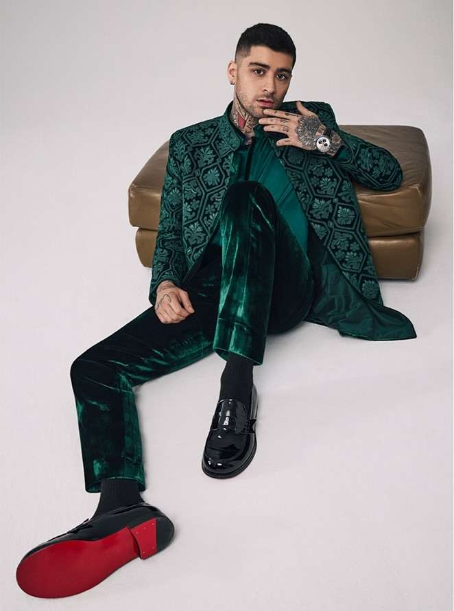 Adult male, Zayn Malik, poses seated in a green patterned jacket and green velvet pants indoors with an ottoman in the background.