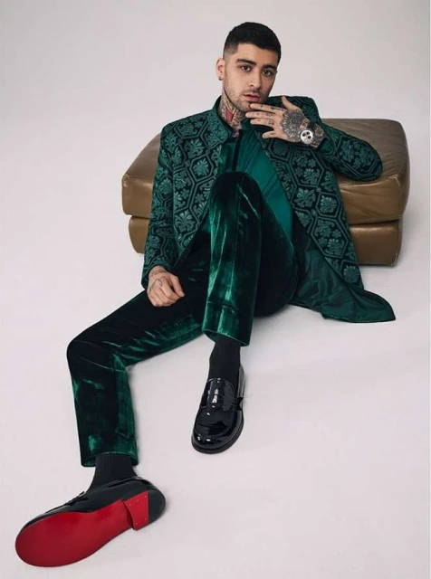 Adult male, Zayn Malik, poses seated in a green patterned jacket and green velvet pants indoors with an ottoman in the background.