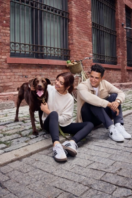 Young woman pets a brown Labrador dog sitting beside a man on cobblestone street, with a bicycle and brick building in the background.