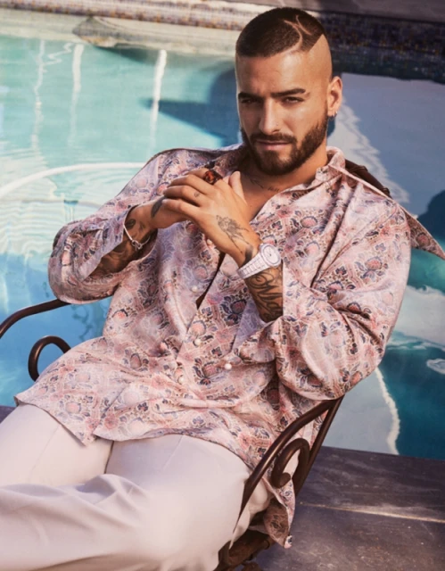 A handsome man wearing a patterned silk shirt is seated by a pool, showing off his tattoos, watch, and decorative haircut.