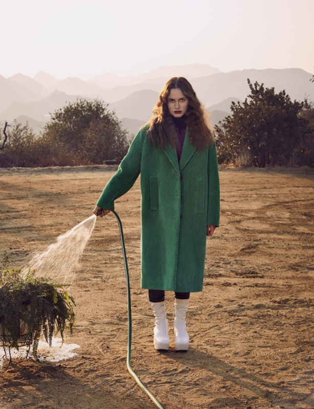 Model in a green coat holding a garden hose watering a plant outdoors with mountains in the background