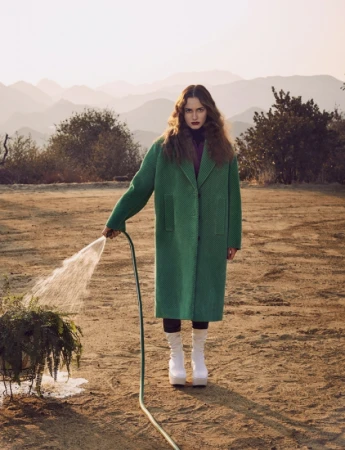 Model in a green coat holding a garden hose watering a plant outdoors with mountains in the background