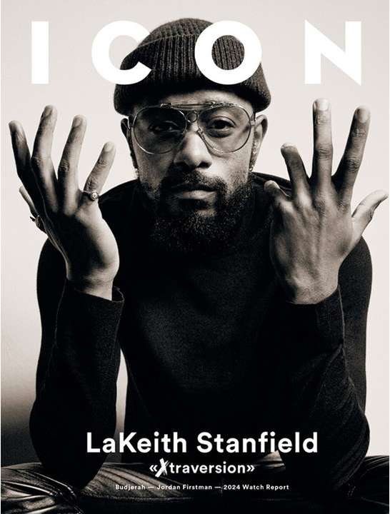 Black and white portrait of LaKeith Stanfield on magazine cover, wearing a beanie and round glasses, raising his hands in a studio setting.