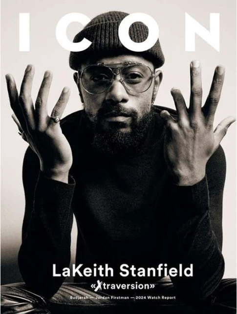 Black and white portrait of LaKeith Stanfield on magazine cover, wearing a beanie and round glasses, raising his hands in a studio setting.