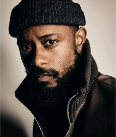 Portrait of a Black man with a beard and dark beanie wearing a zipped jacket against a plain light background