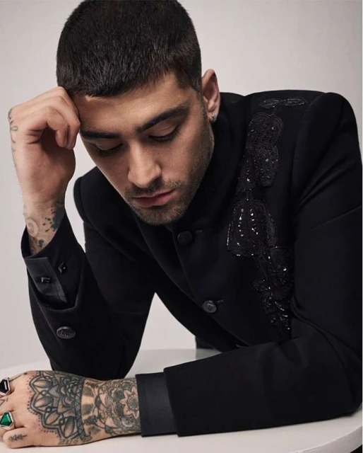 Close-up portrait of adult male in black suit jacket with embellishments, leaning his head on his hand with tattoos on his arms.