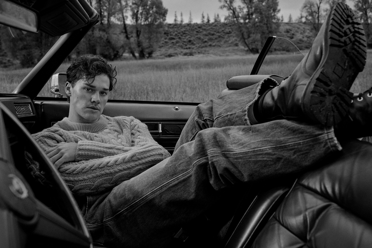 Monochrome shot of a young adult male sitting in a car with his boots up on the headrest in front of a grassy field.