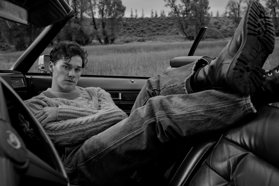 Monochrome shot of a young adult male sitting in a car with his boots up on the headrest in front of a grassy field.