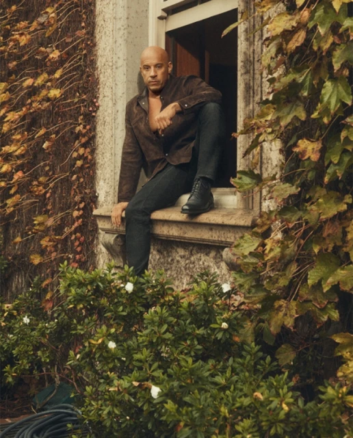 Bald adult man with a brown open button-down shirt and dark denim jeans sitting on a windowsill surrounded by ivy and lush green foliage.