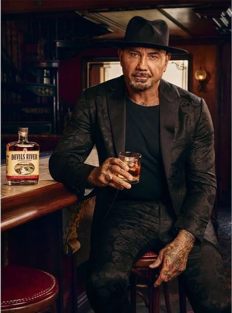 Actor Dave Bautista in a dark floral suit holding a glass of whiskey with a Devils River bottle on a bar, against a dark background.