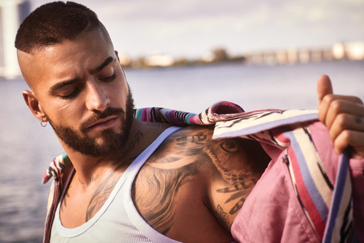Close-up of a man with short dark hair and tattoos, wearing a white tank top and a patterned jacket, standing outdoors near a body of water.