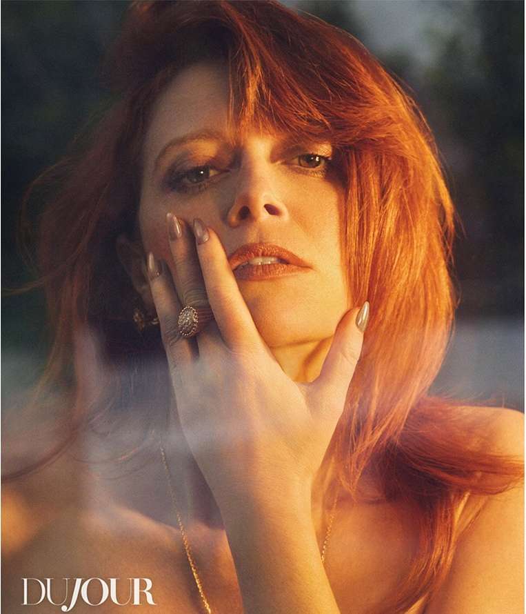 Close-up portrait of a woman with red hair and a golden glow on her face, wearing a ring and necklace, with the 'DuJour' logo.