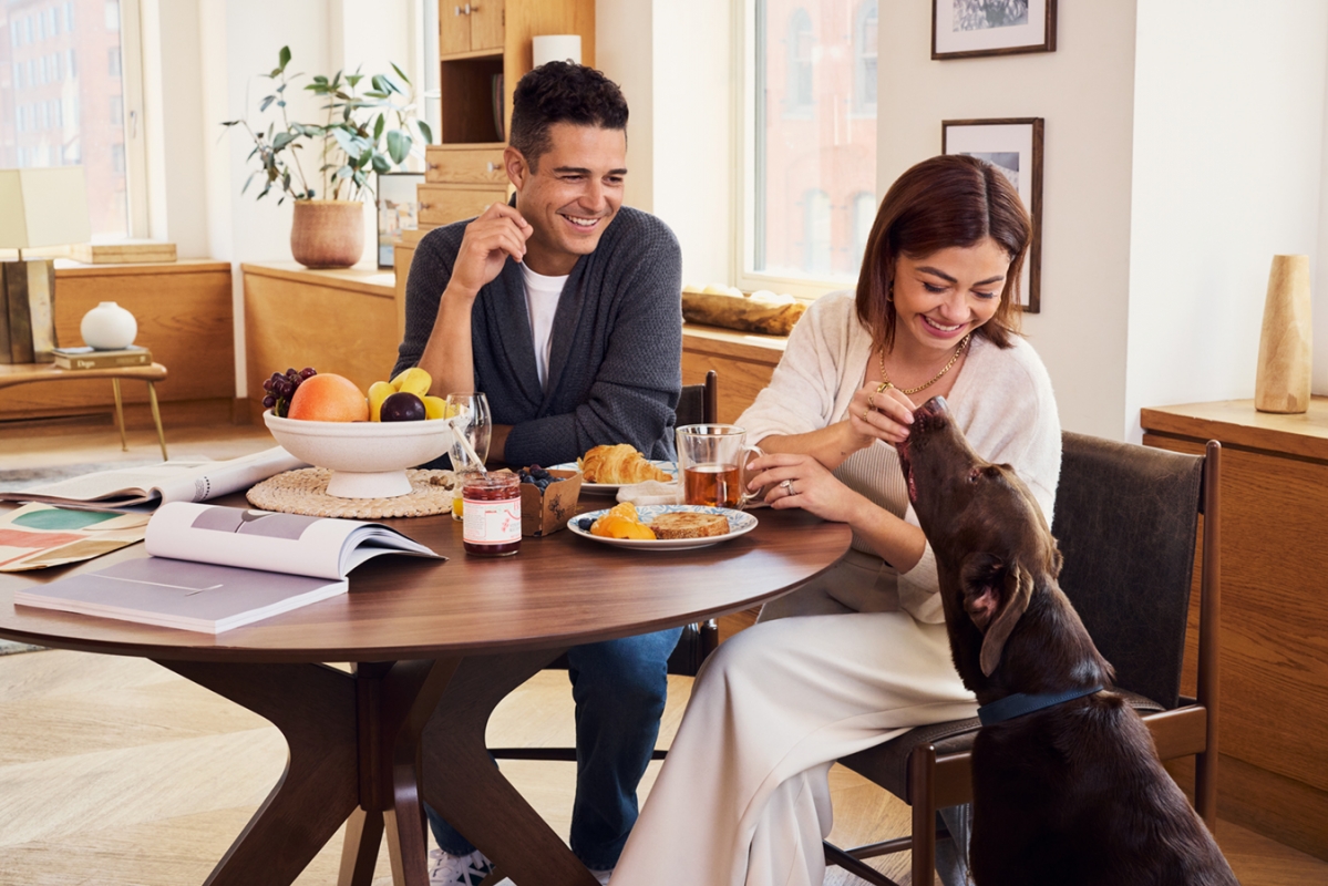 A smiling couple enjoys a meal at a round wooden table with their brown dog, featuring a fruit bowl, croissants, and warm natural lighting.