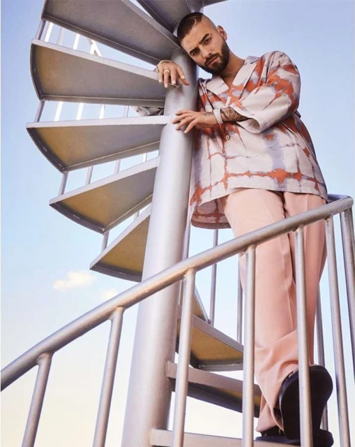 Adult male leaning on a spiral staircase, wearing a printed shirt and pink trousers against a sky background.