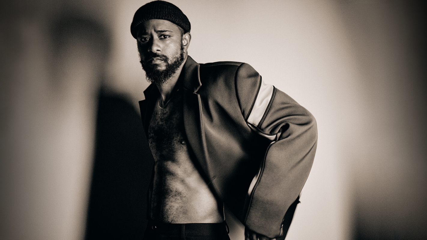 Black man wearing a beanie and a structured jacket over a shirtless torso against a gradient background.