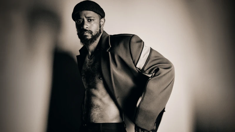 Black man wearing a beanie and a structured jacket over a shirtless torso against a gradient background.